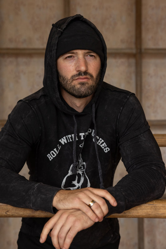 Man wearing a black hoodie with text and graphics, sitting against a wooden paneled wall.