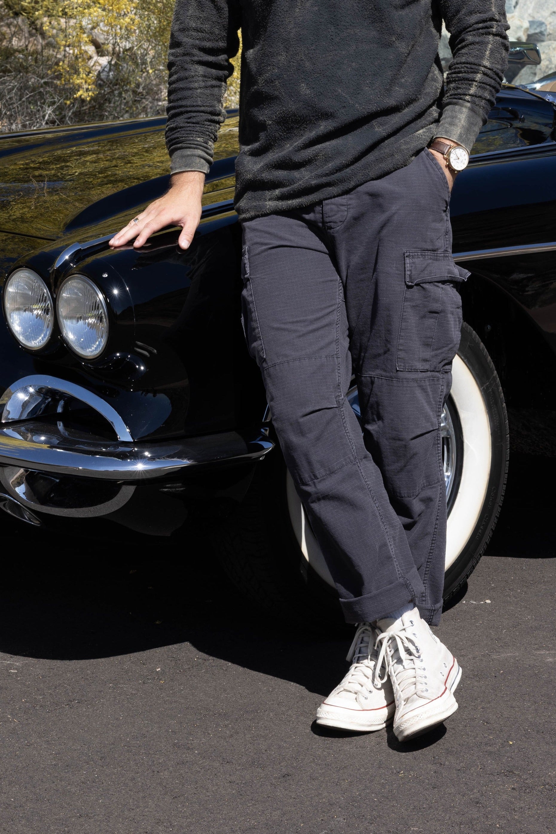 Man leaning against a classic black car in a scenic outdoor setting with trees and rocks.