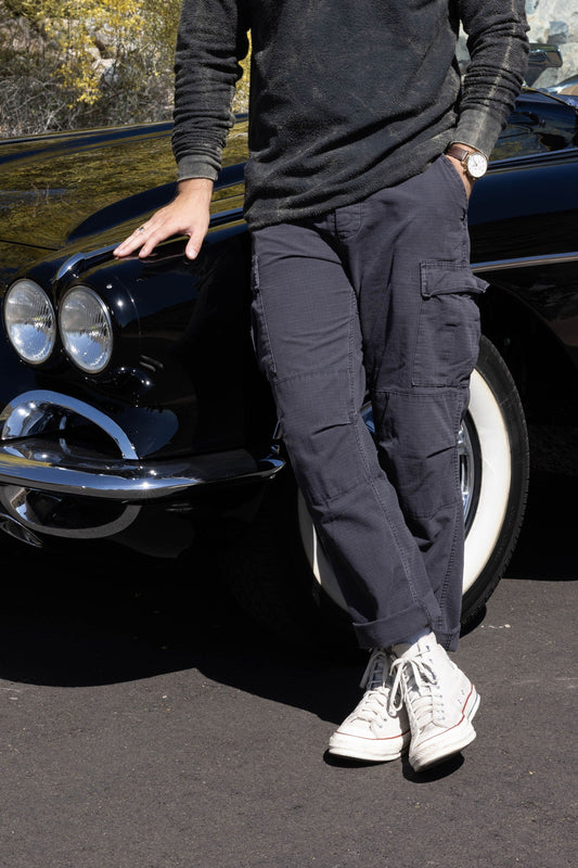 Man leaning against a classic black car in a scenic outdoor setting with trees and rocks.